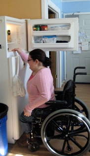 This image shows a woman in a wheelchair in front of her refrigerator attempting to reach into her freezer. The freezer is located above the fridge making it nearly impossible for her to access anything inside because the freezer is too high. 